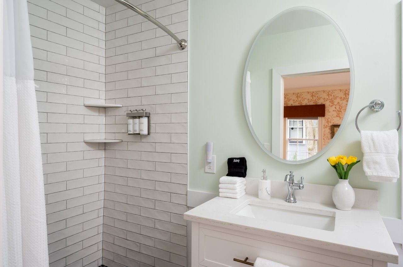 A bright bathroom with a white subway-tile shower, oval mirror over a white vanity, and a vase of yellow tulips.