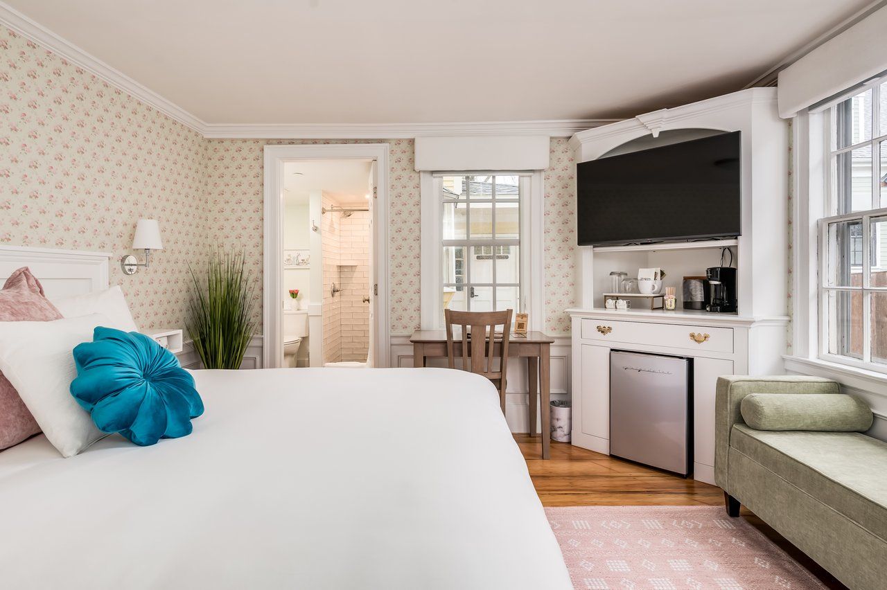 A well-lit hotel bedroom with a large bed, patterned wallpaper, a television, a desk, a mini-fridge, and an en-suite bathroom.