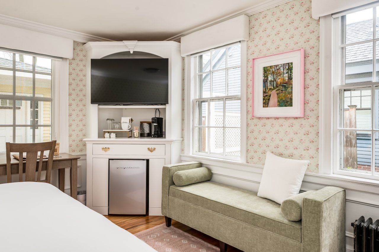 A cozy hotel room corner with a flat-screen TV on a white stand, a mini-fridge, a sage green bench, and floral wallpaper.