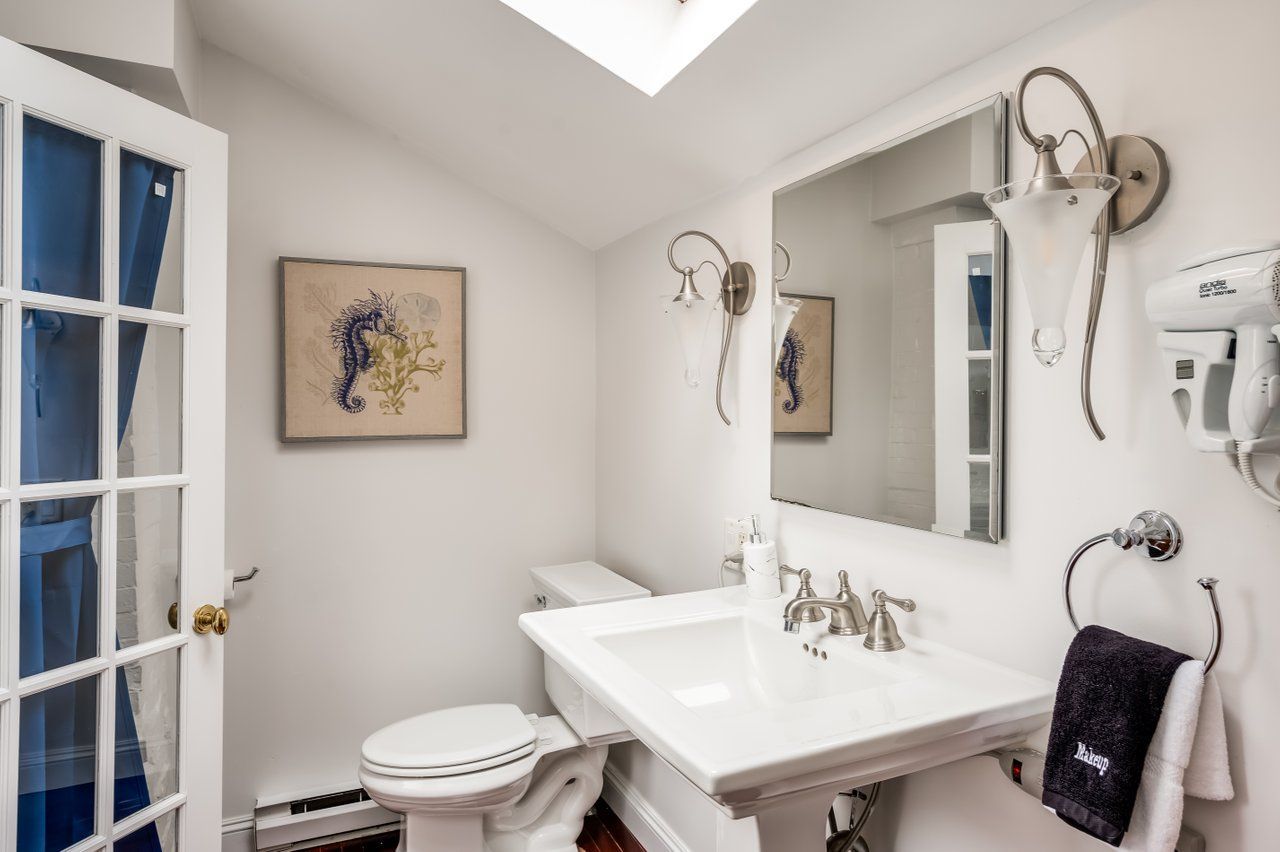 A bright bathroom featuring a white pedestal sink, toilet, rectangular mirror, two wall sconces, and a framed artwork.