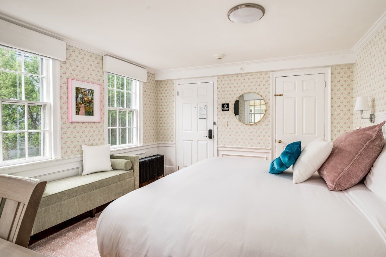 A clean hotel room with a large bed, light patterned wallpaper, windows, a bench, and two white doors.