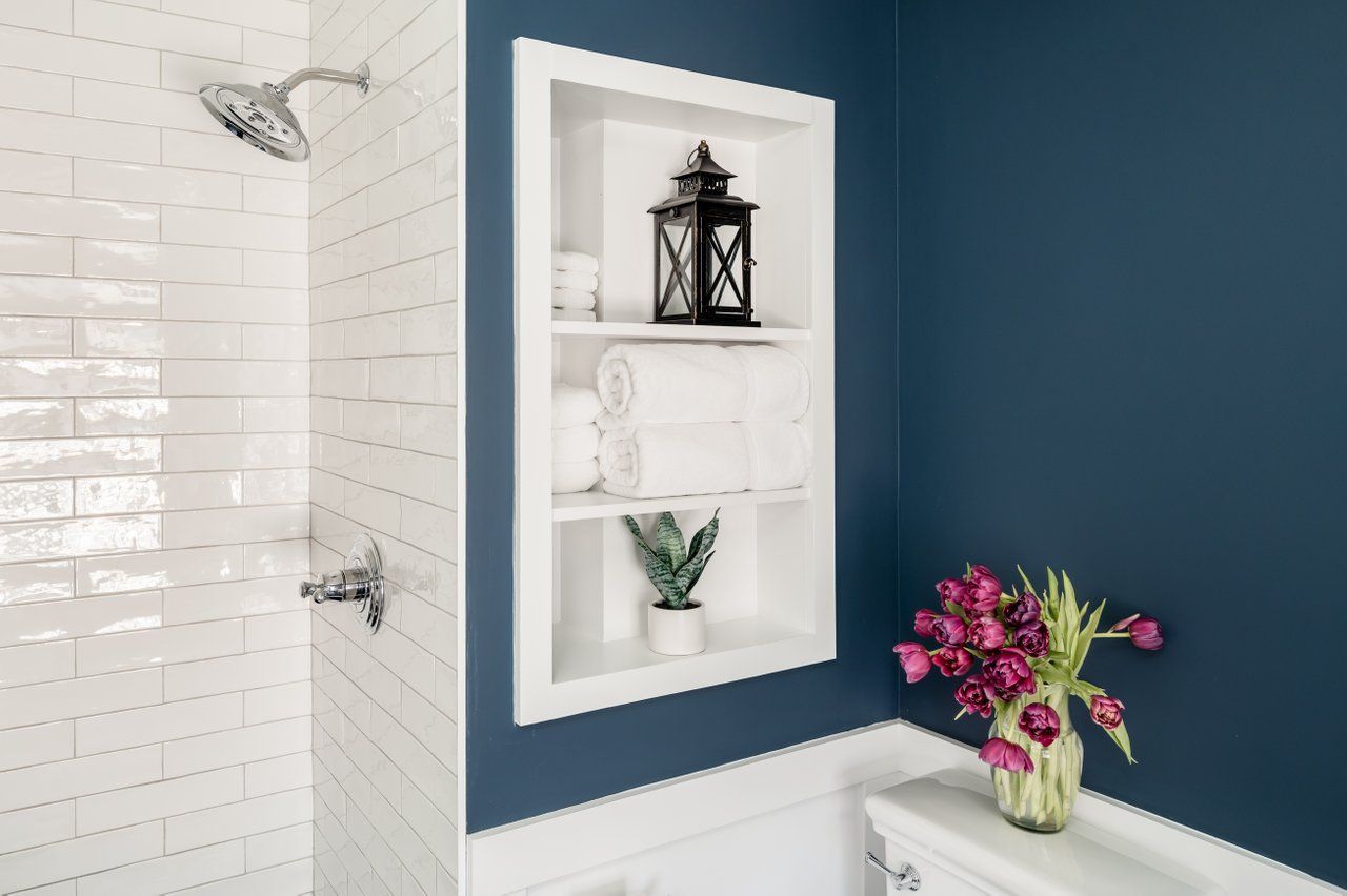 A built-in white shelf set into a deep blue wall next to a white tiled shower, with towels, decor, and a vase of flowers.