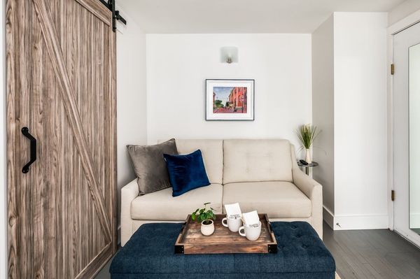 Interior view of a modern hotel suite featuring a beige sofa, blue-accented bedroom, and a glass-paneled exterior door.