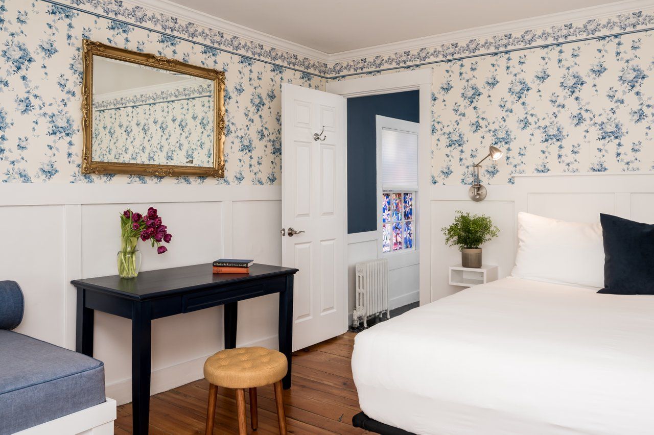 A bedroom with floral wallpaper, a white bed, a dark desk with a mirror above it, and an open doorway to another room.