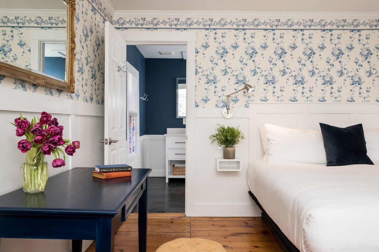 A bedroom with floral wallpaper and white wainscoting, featuring a blue desk, a bed, and a door leading to a blue bathroom.