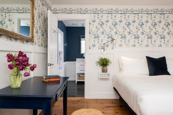 A bedroom with floral wallpaper, a dark blue desk with purple flowers, a bed, and an open doorway to a blue bathroom.