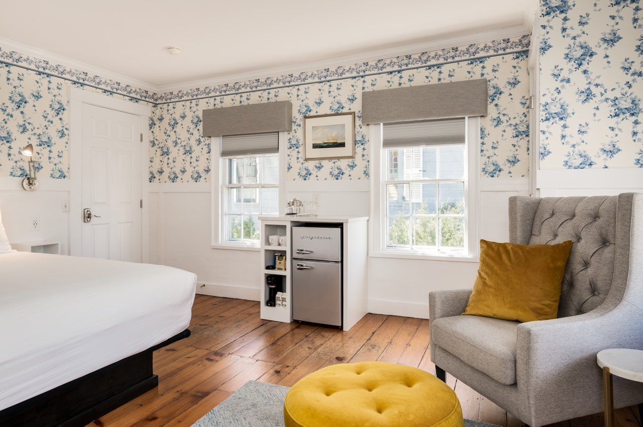A hotel room with a bed, a gray armchair with a yellow pillow, a yellow ottoman, and blue-and-white floral wallpaper.