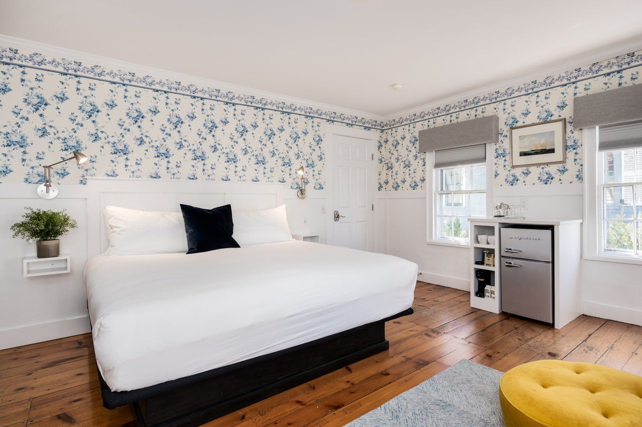 A hotel room with a large bed, blue floral wallpaper, white wainscoting, a mini-fridge, and a round yellow ottoman.