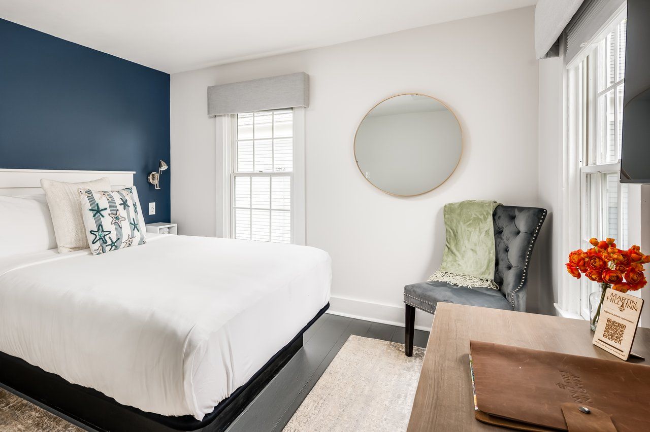A hotel room featuring a queen bed with a blue accent wall, a gray tufted armchair, round mirror, and a wooden desk.