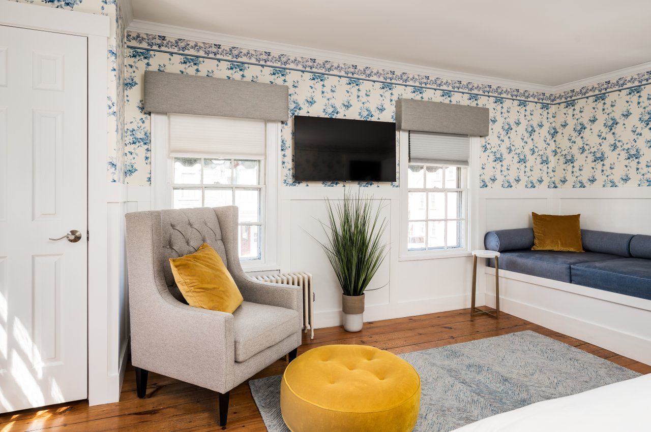 A cozy sitting area with blue floral wallpaper, a grey armchair, a yellow ottoman, and a built-in blue window seat.