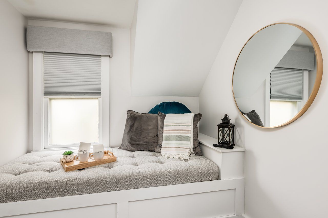 A window seat with gray tufted cushions, pillows, and a serving tray under a round mirror in a white-walled room.