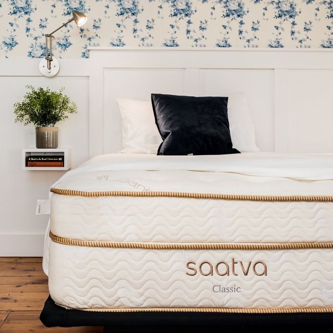 A Saatva Classic mattress on a bed frame against a white headboard and blue floral wallpaper with a wall-mounted lamp.