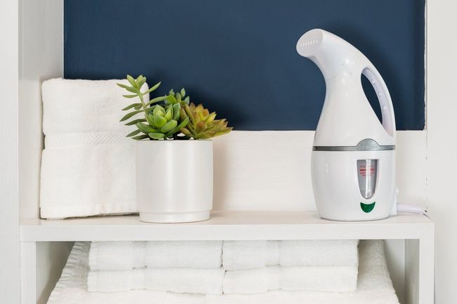 A white handheld garment steamer, a small potted succulent, and a folded white towel on a white shelf against a blue wall.