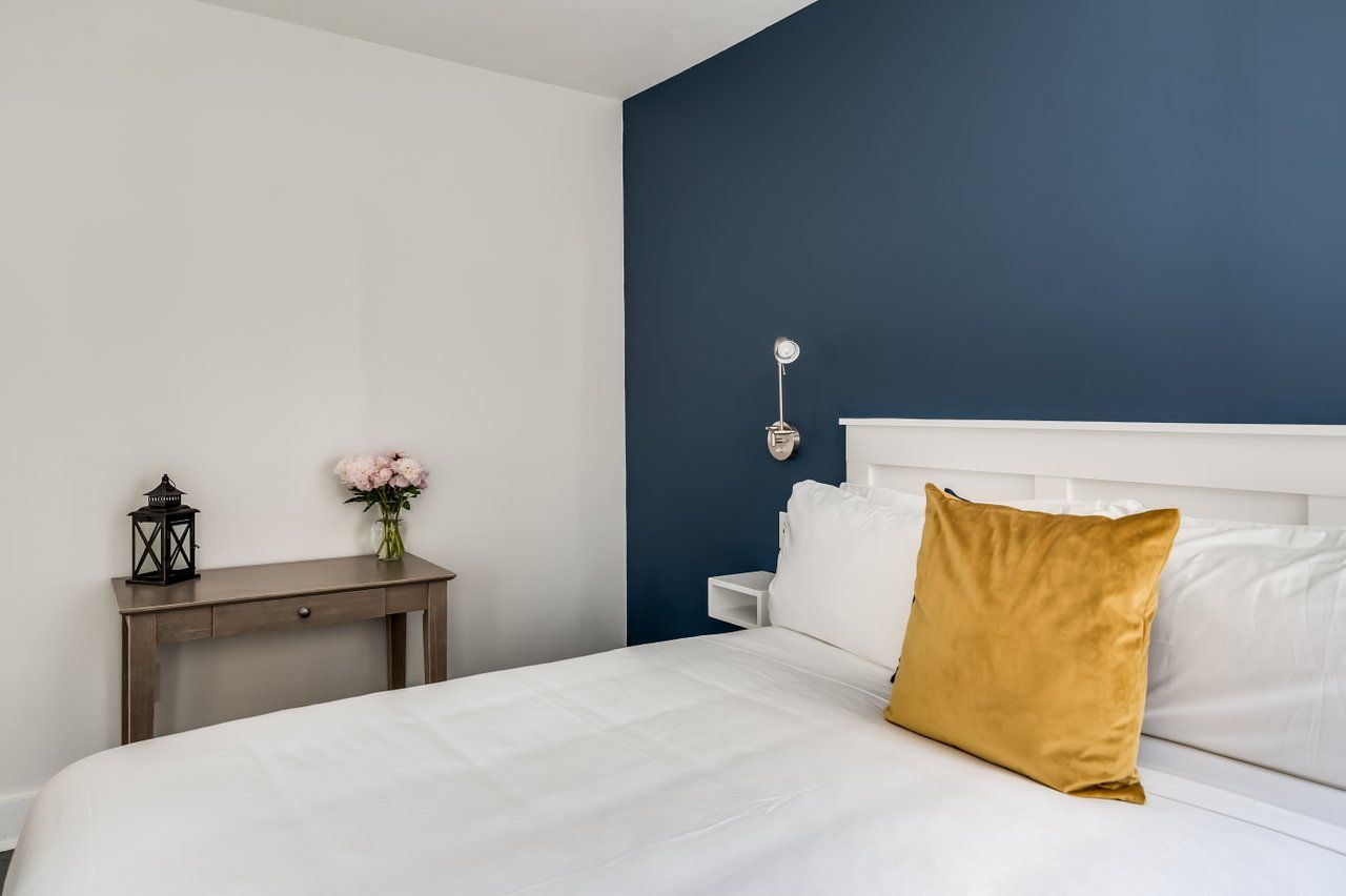 A bedroom with white bedding, a gold pillow, a small wooden desk, and a dark blue accent wall.