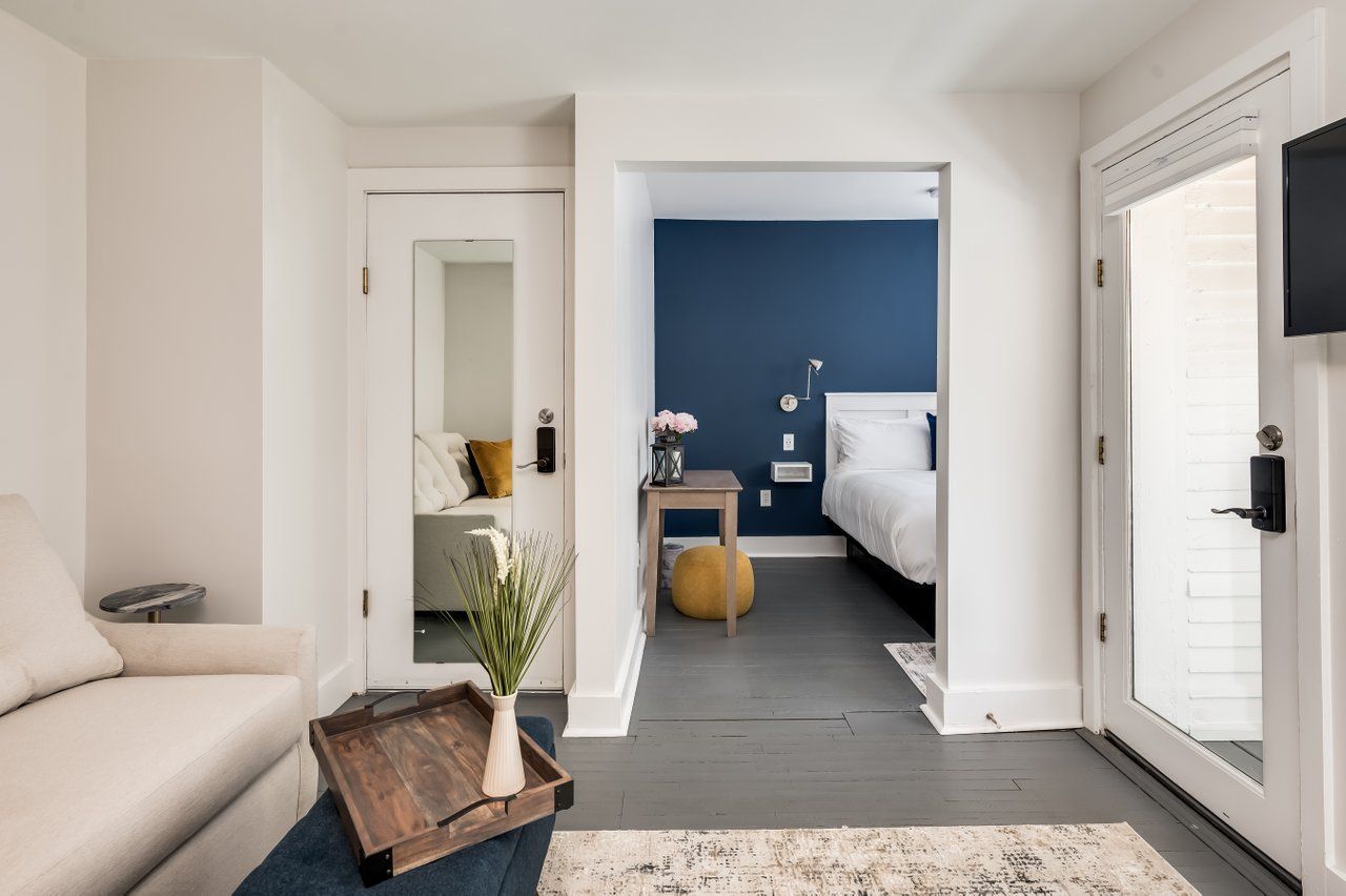 A view from a living area with a sofa into a bedroom with a blue accent wall, white bed, and side table.