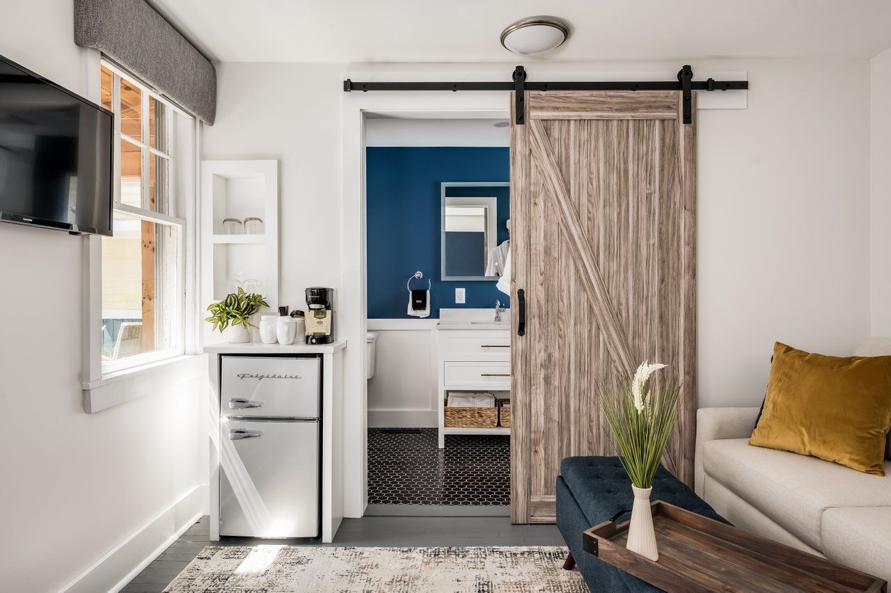 A cozy room with a mini fridge, wooden sliding barn door leading to a blue-walled bathroom, and a sofa with gold cushion.
