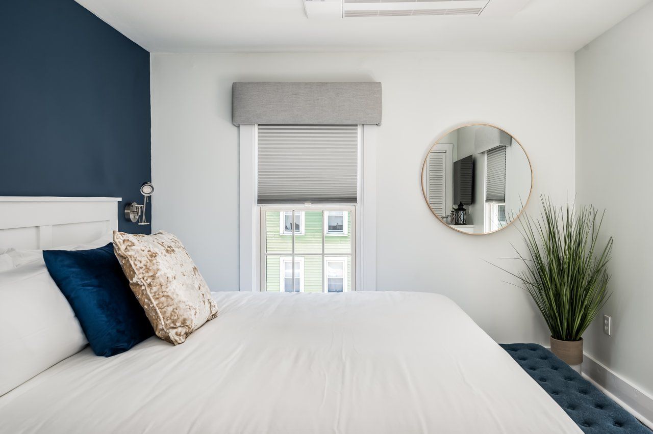 A modern bedroom with a white bed, navy accent wall, grey window shade, circular mirror, and a green potted plant.