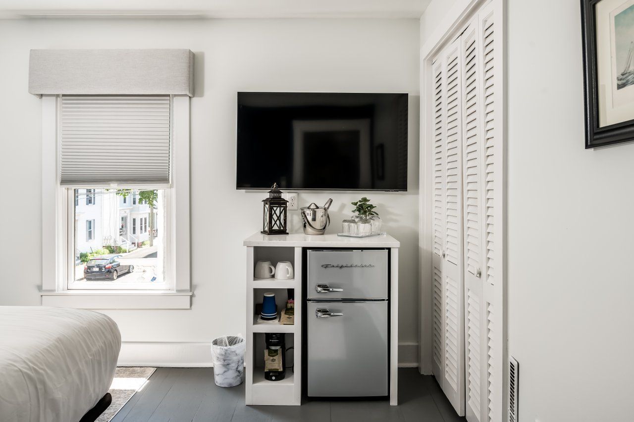 A modern bedroom corner with a mounted TV above a white cabinet featuring a small silver mini-fridge and coffee supplies.