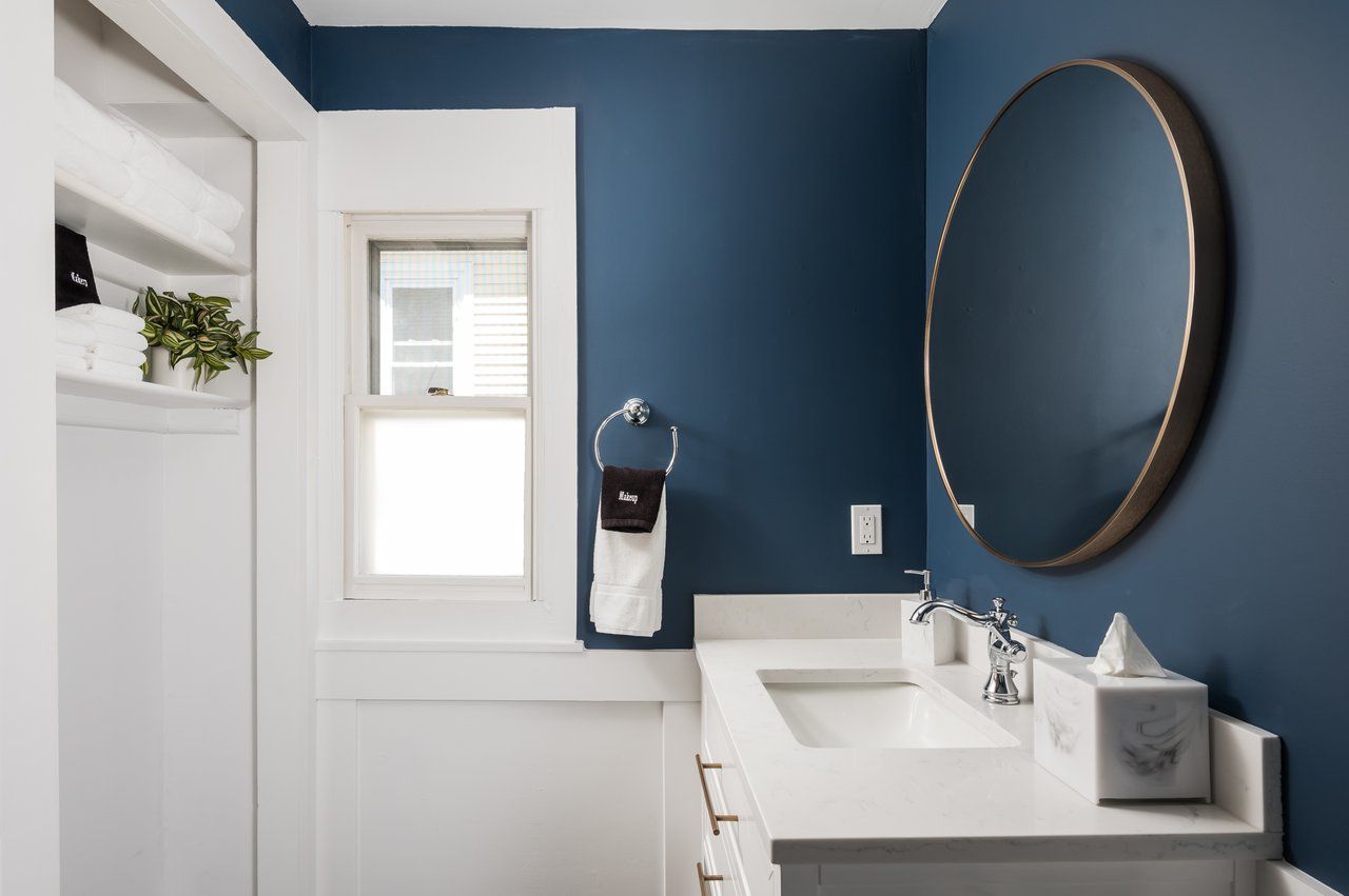 Modern bathroom with deep blue walls, white trim, a circular vanity mirror, and a white countertop sink.