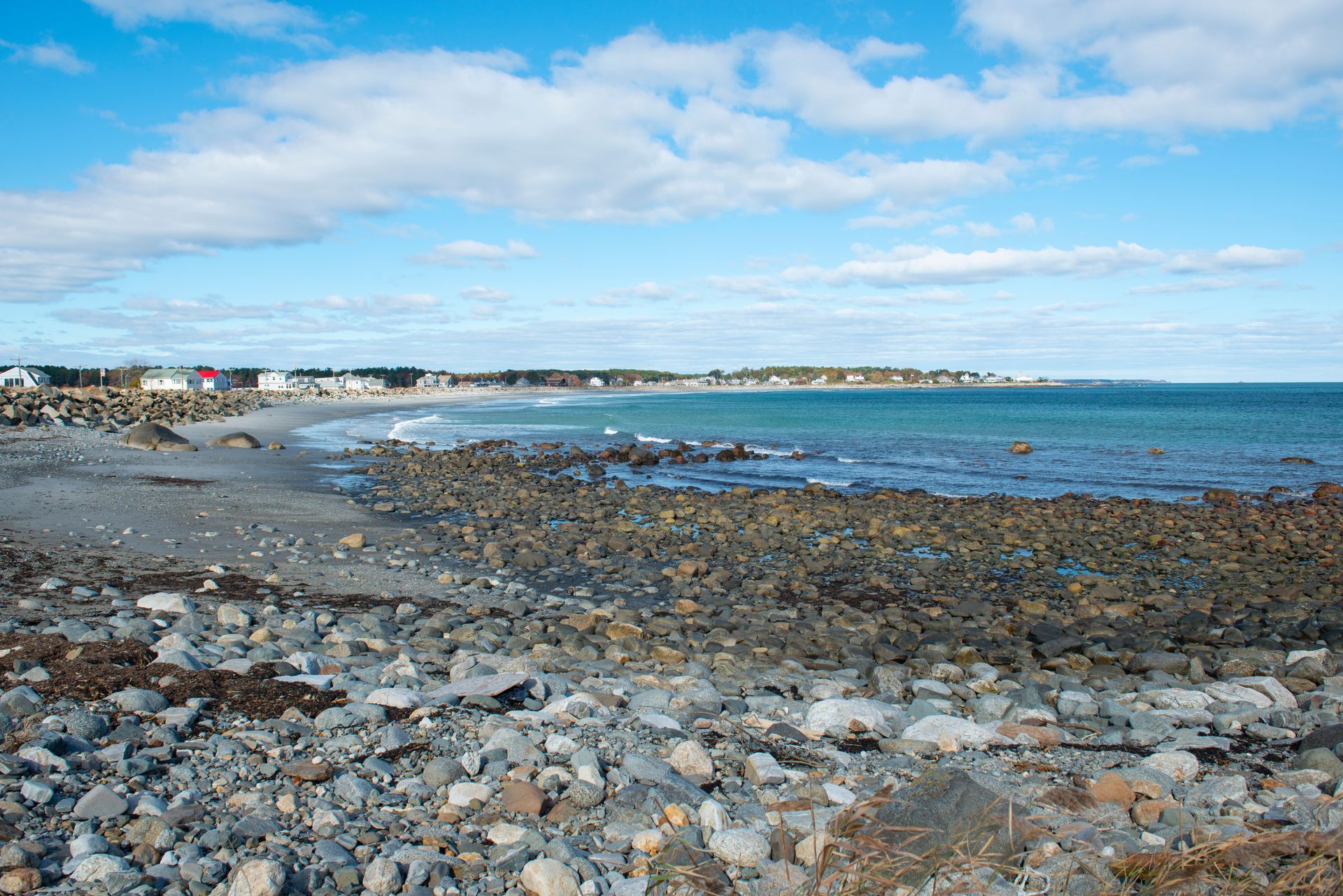 Rocky shoreline with a beach, coastal houses in the distance, and blue ocean under a partly cloudy sky.