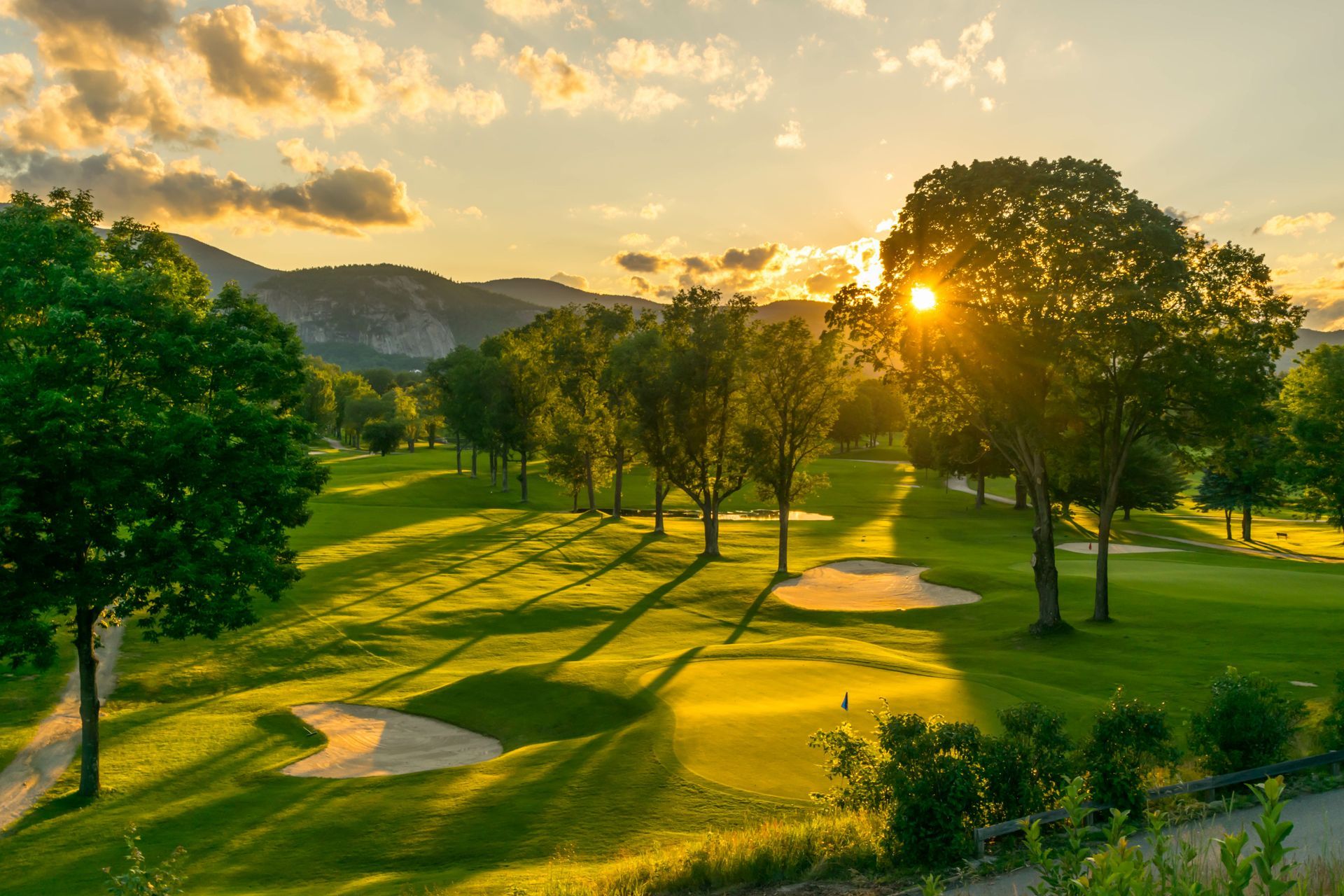 A sunlit golf course at golden hour with lush green fairways, sand traps, and silhouetted trees against a mountain range.