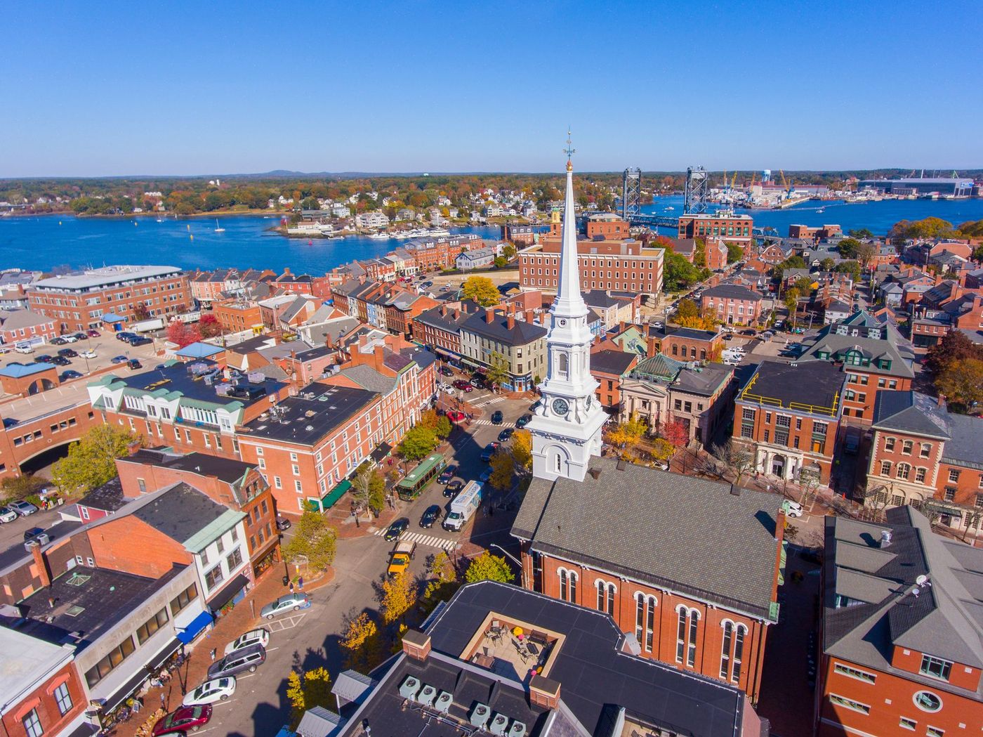 An aerial view of a New England town with a prominent white church steeple overlooking buildings and a nearby harbor.
