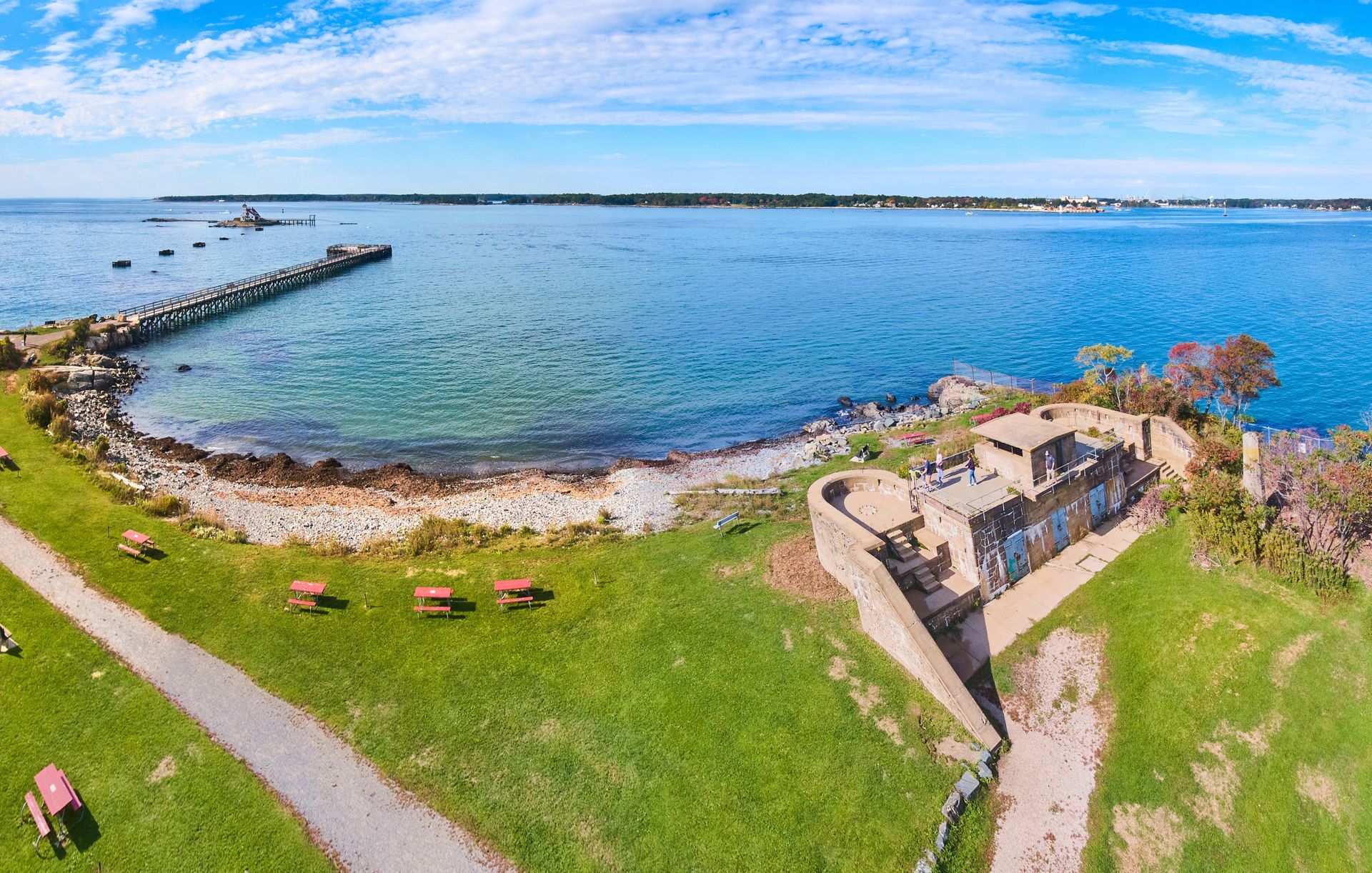 Aerial view of a coastal park with a concrete bunker ruin, picnic tables on grass, and a stone pier extending into the sea.