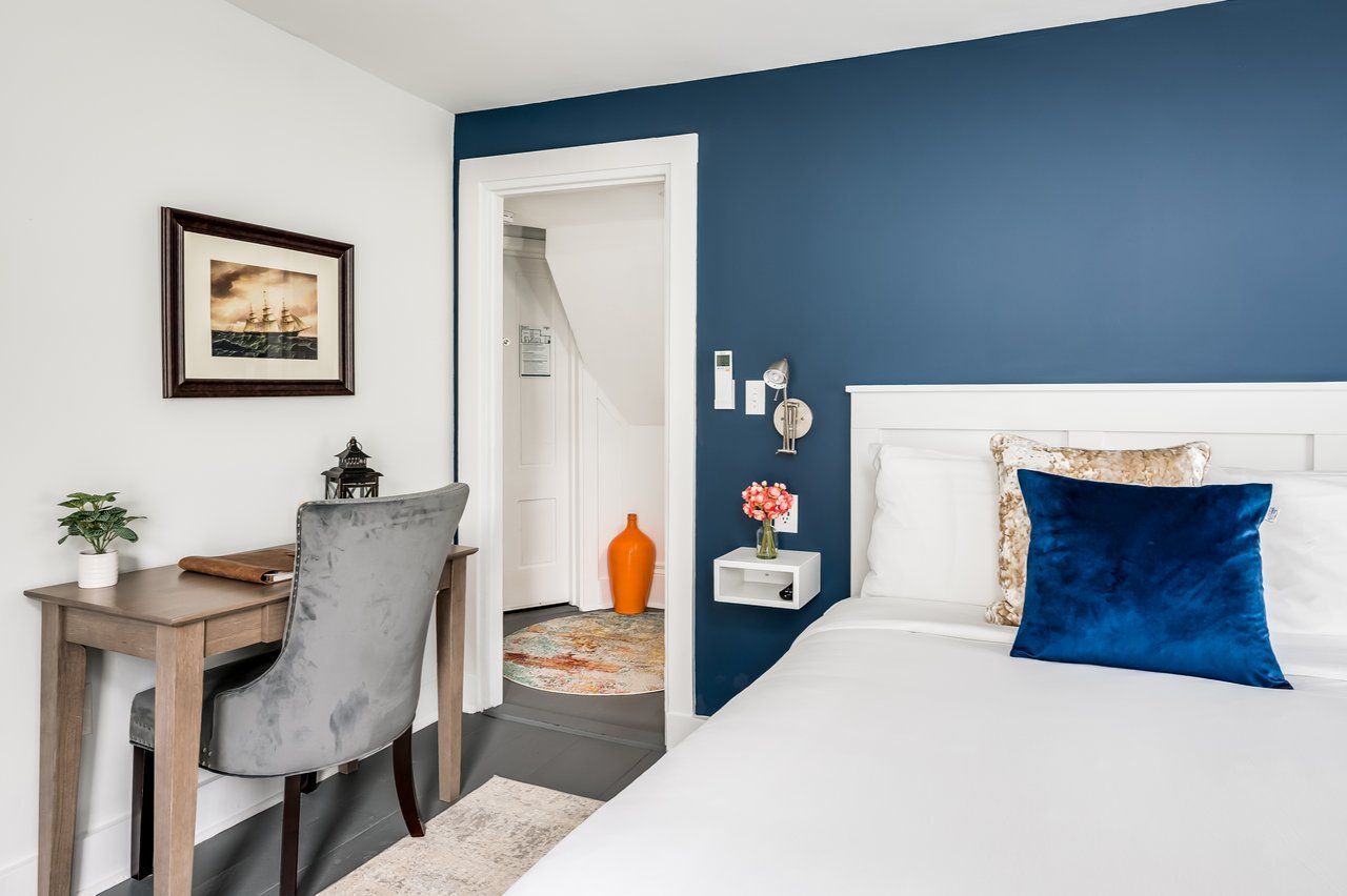 A bedroom with a bed featuring a blue pillow, a desk with a grey chair, and a doorway leading to a small hallway.