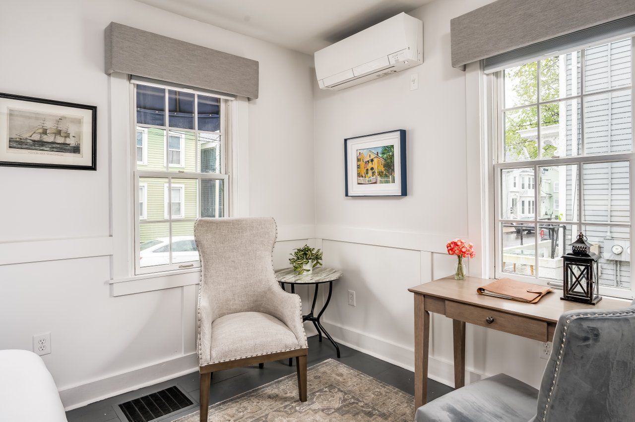 A cozy corner with a white armchair, a small round side table, a wooden desk with a chair, and a wall-mounted AC unit.
