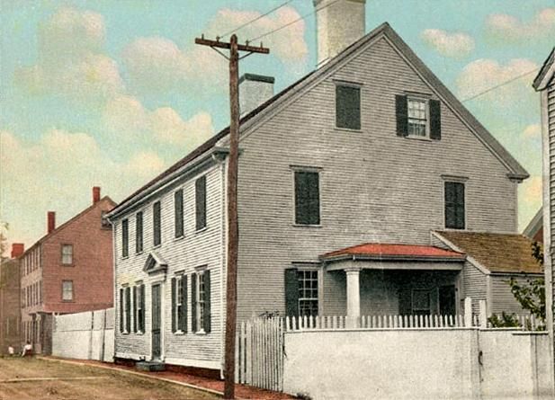 A two-story white clapboard house with a small front porch and a picket fence, set next to a red brick building.