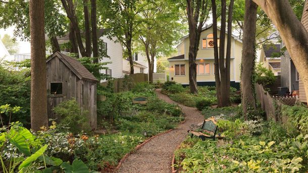 A dirt path winds through a lush, tree-shaded garden toward a light-colored house with a small wooden shed on the left.