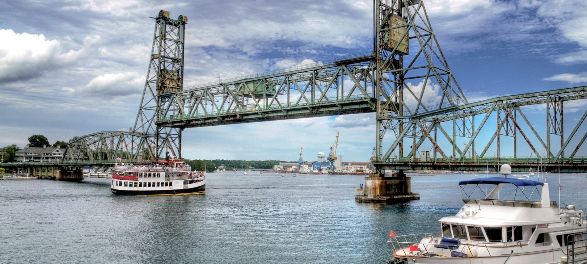A blue vertical-lift bridge spans a river with a passenger boat and a white yacht cruising below under a cloudy sky.