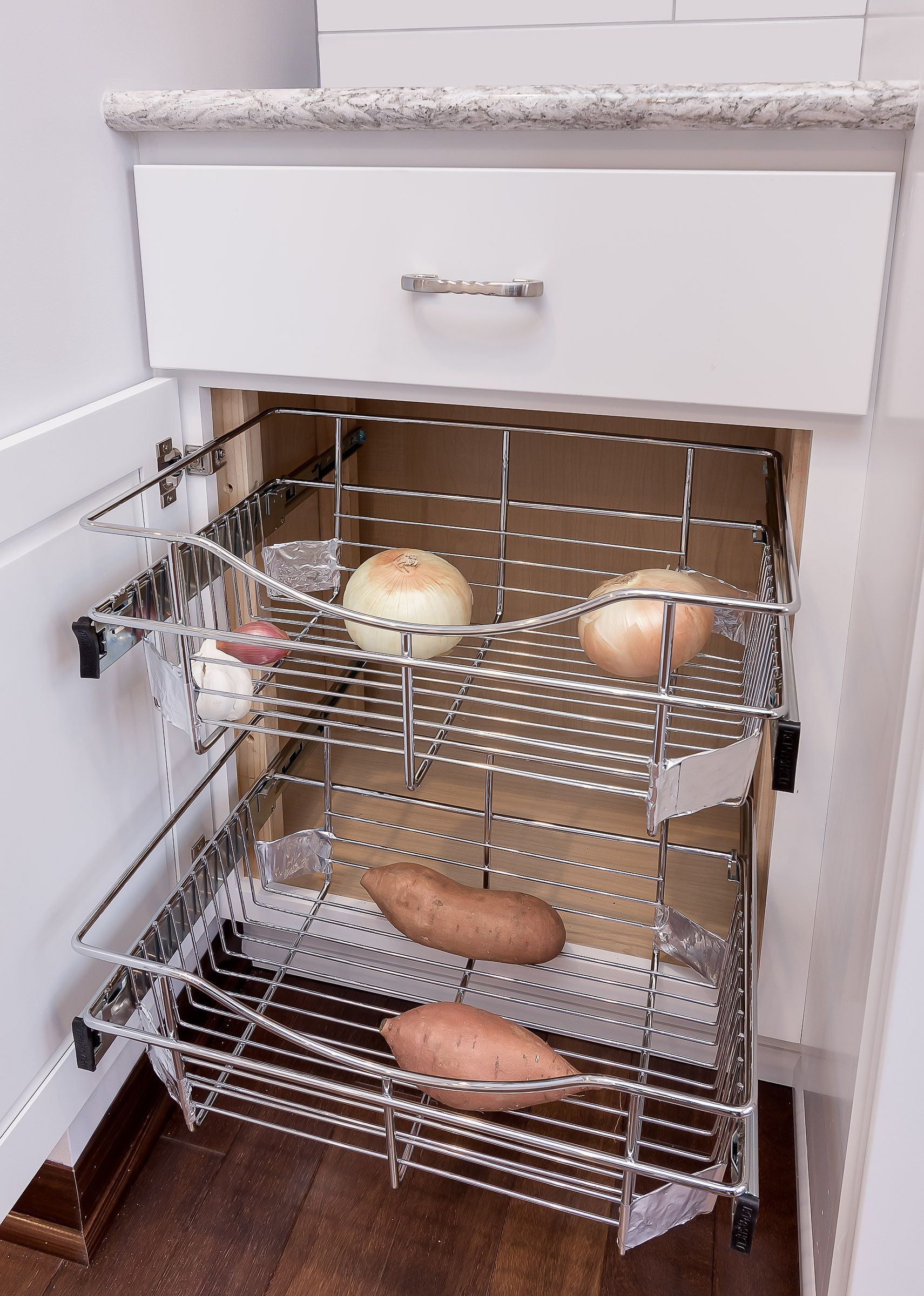 A kitchen cabinet with pull-out wire baskets holding onions, sweet potatoes, and garlic.
