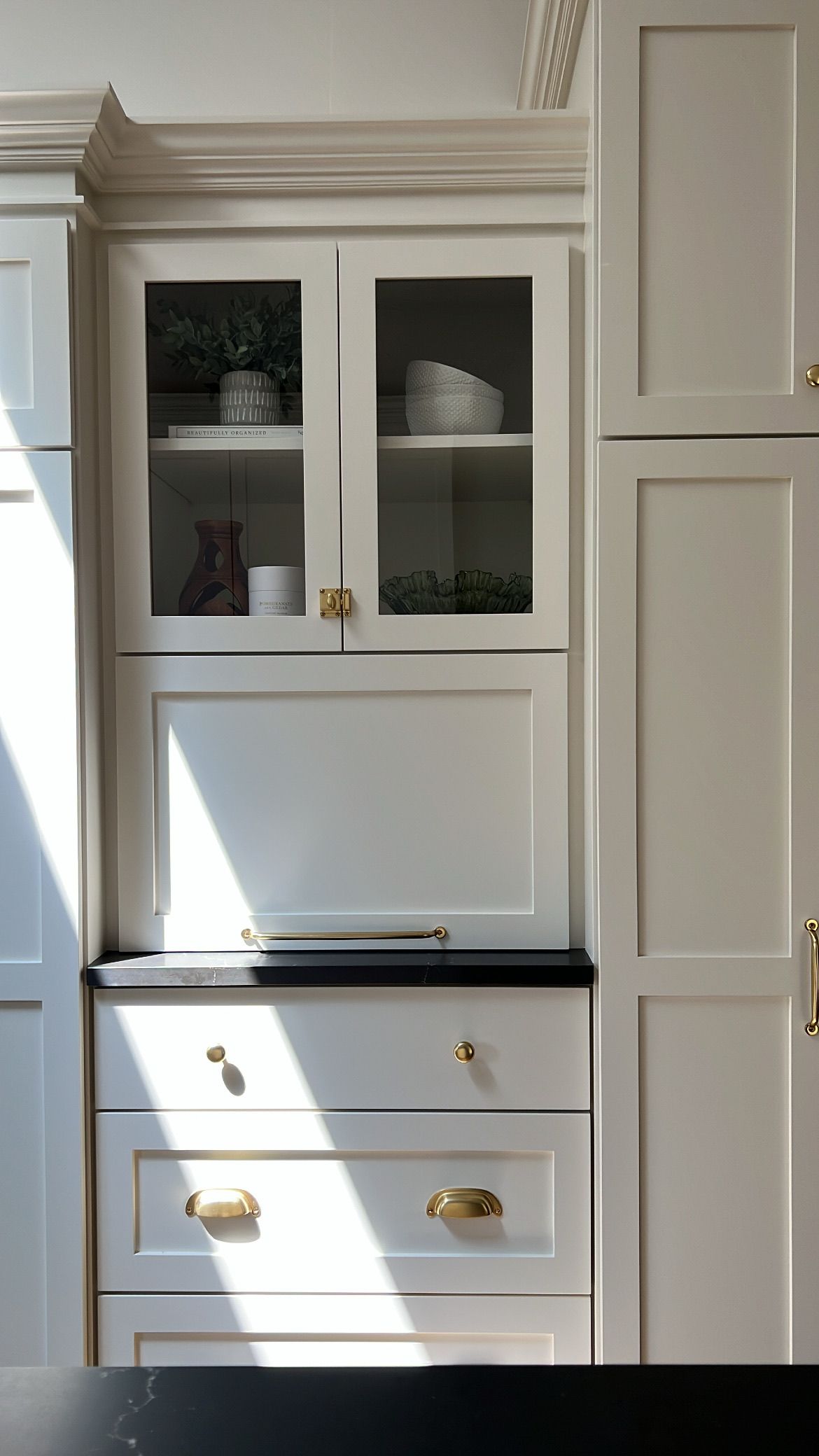 White kitchen cabinets with glass-fronted upper cabinet. Black countertop. Gold hardware. Sunlit.