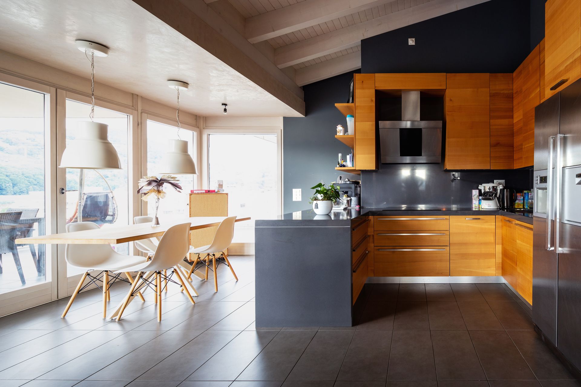 Modern kitchen with wood cabinets, dark countertops, and dining table by large windows.