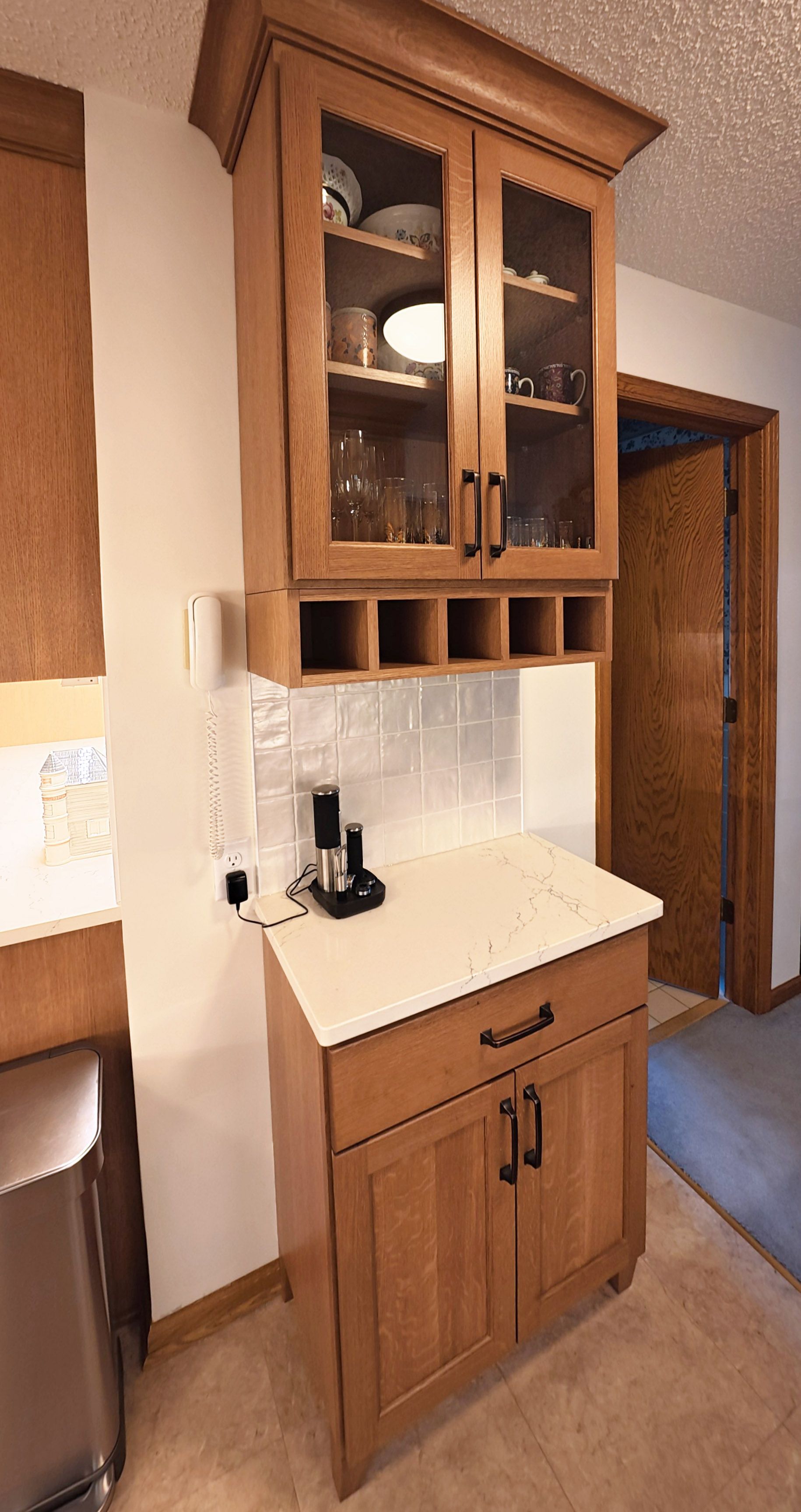 Wooden kitchen cabinet with glass-front upper doors, wine rack, and a lower cabinet against a white tiled wall.
