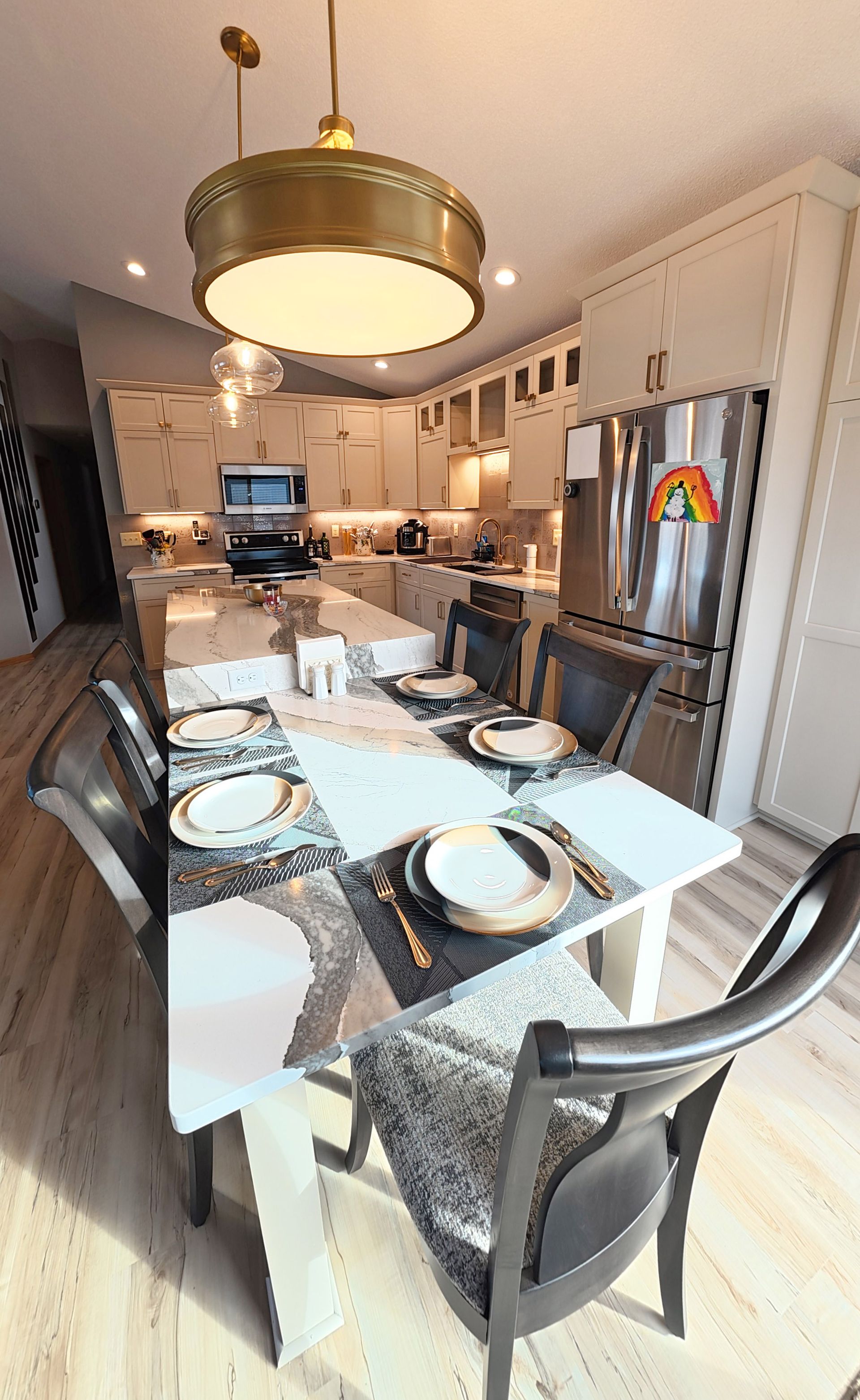 Kitchen with dining table set for a meal. Includes island, cabinets, refrigerator, and pendant lighting.