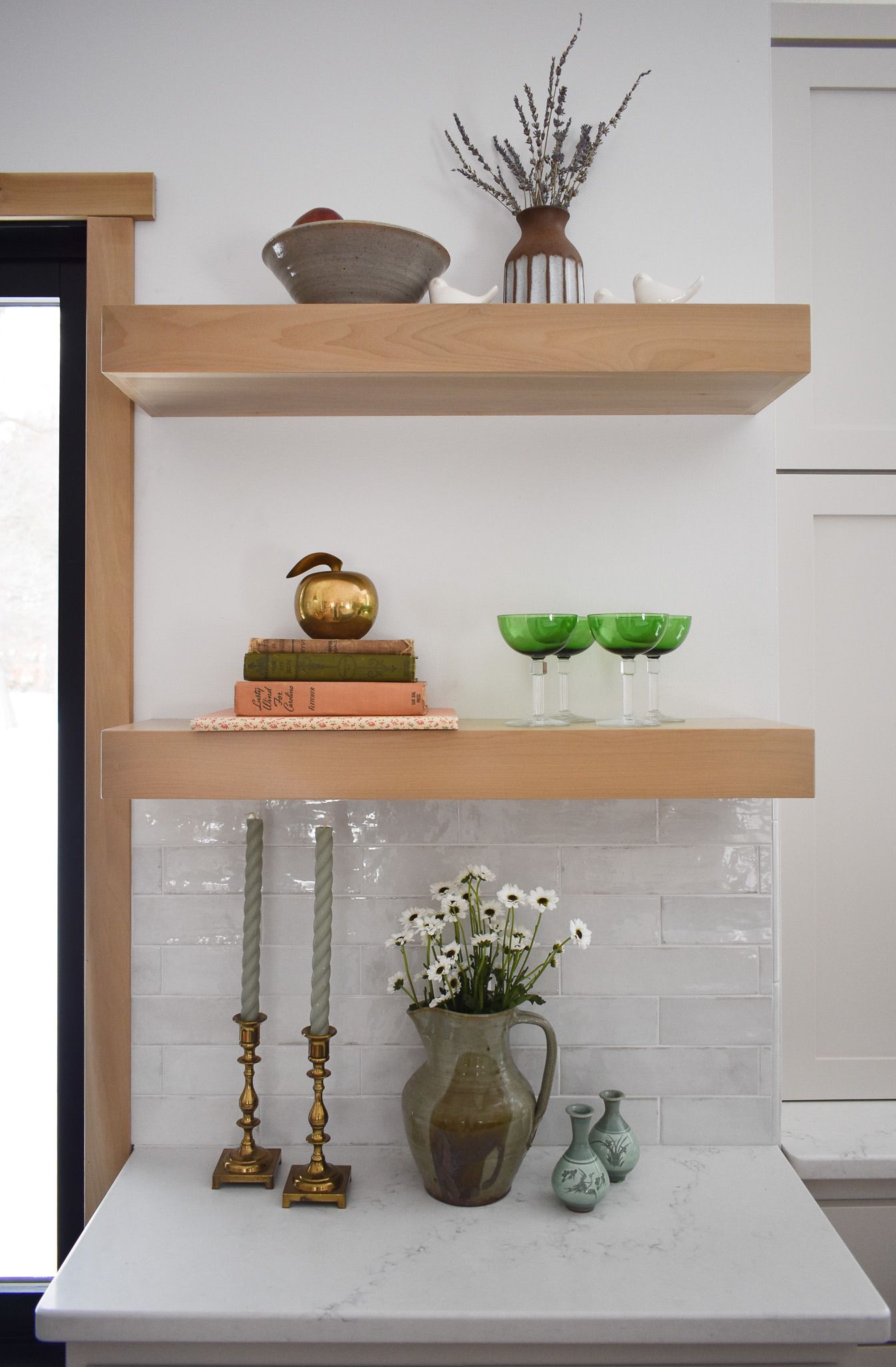 Wooden shelves with decor: flowers, books, candles, glassware. White tile backsplash.