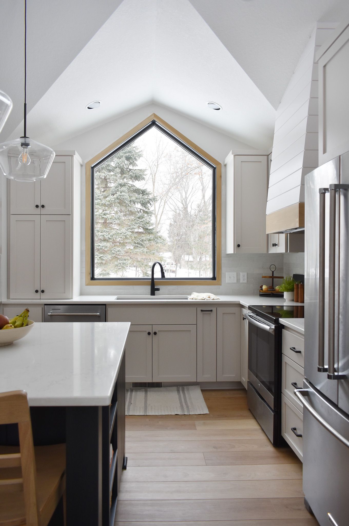 Modern kitchen with white cabinets, dark island, triangle window with a view of snow-covered trees.