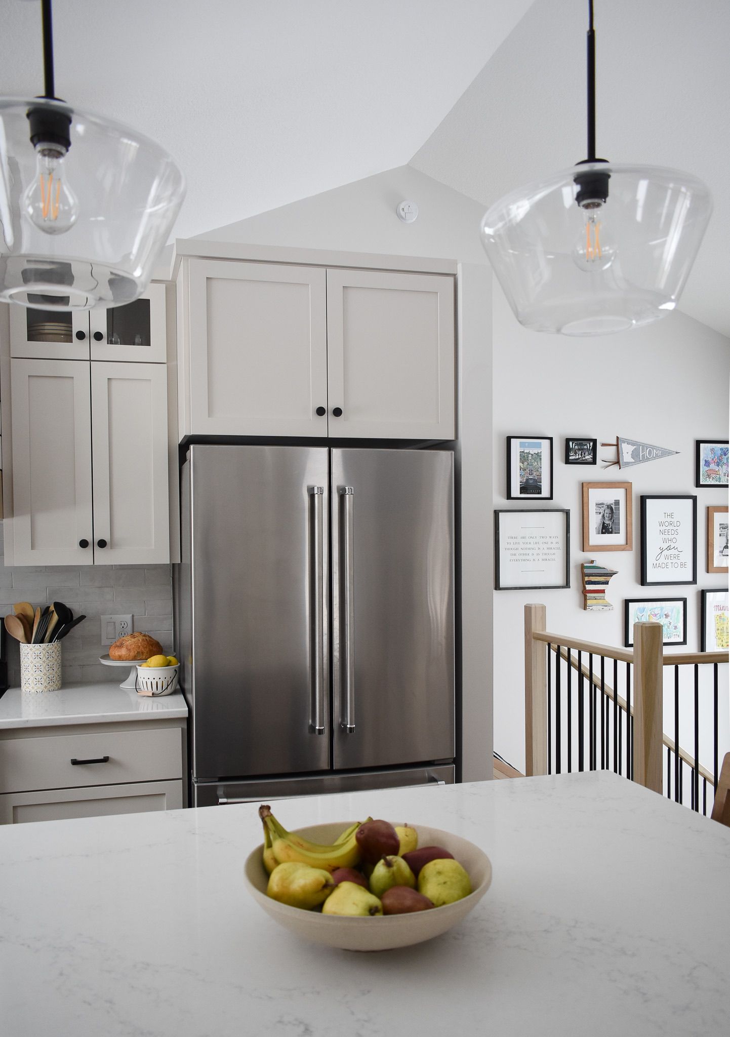 Modern kitchen with stainless steel fridge, light cabinets, glass pendant lights, fruit bowl on the island.