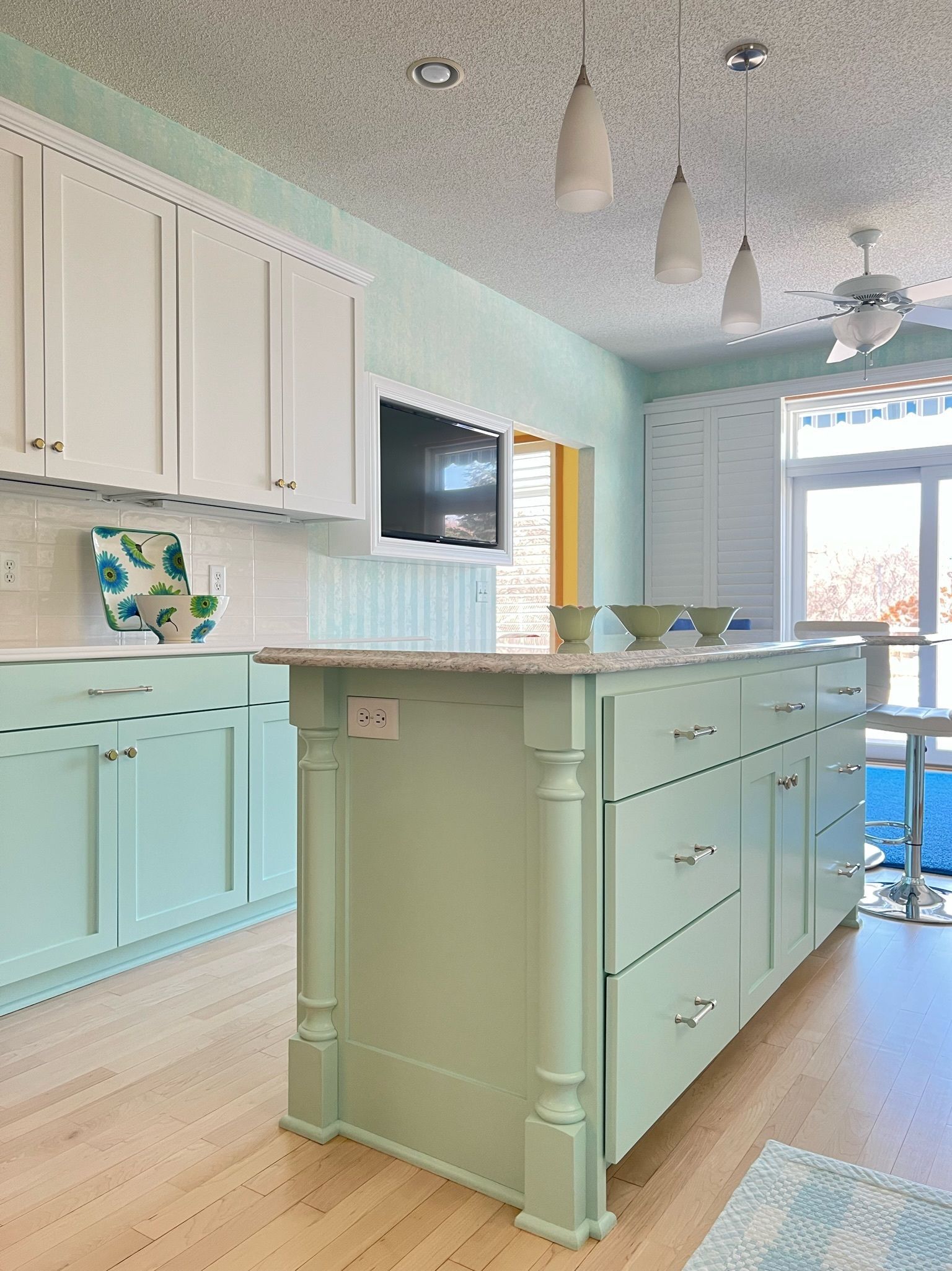 Light green kitchen with island, white cabinets, and wooden floor. A small TV hangs above the island.
