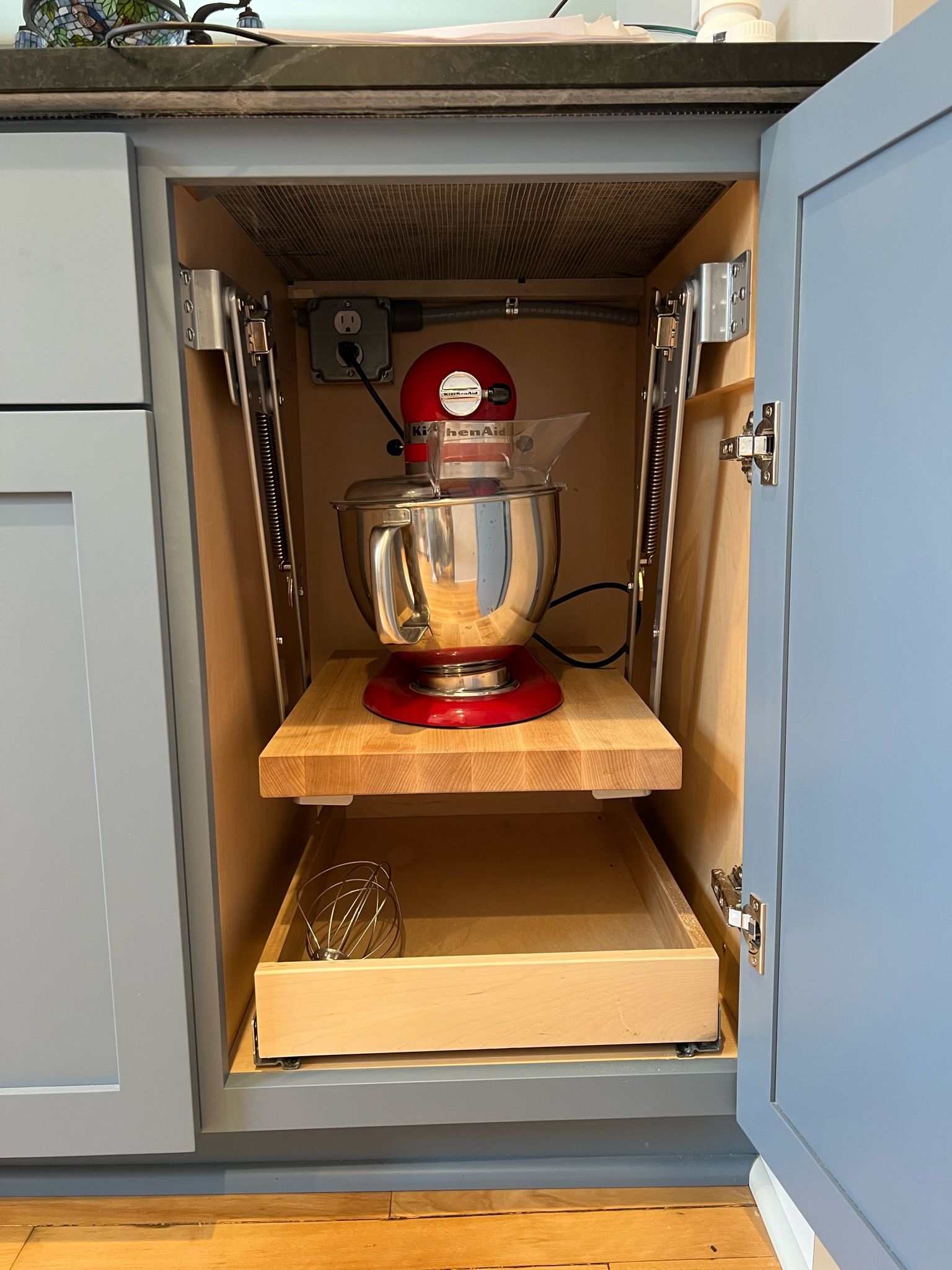 Kitchen cabinet with red stand mixer on a pull-out shelf; blue doors. A lower drawer holds a whisk.