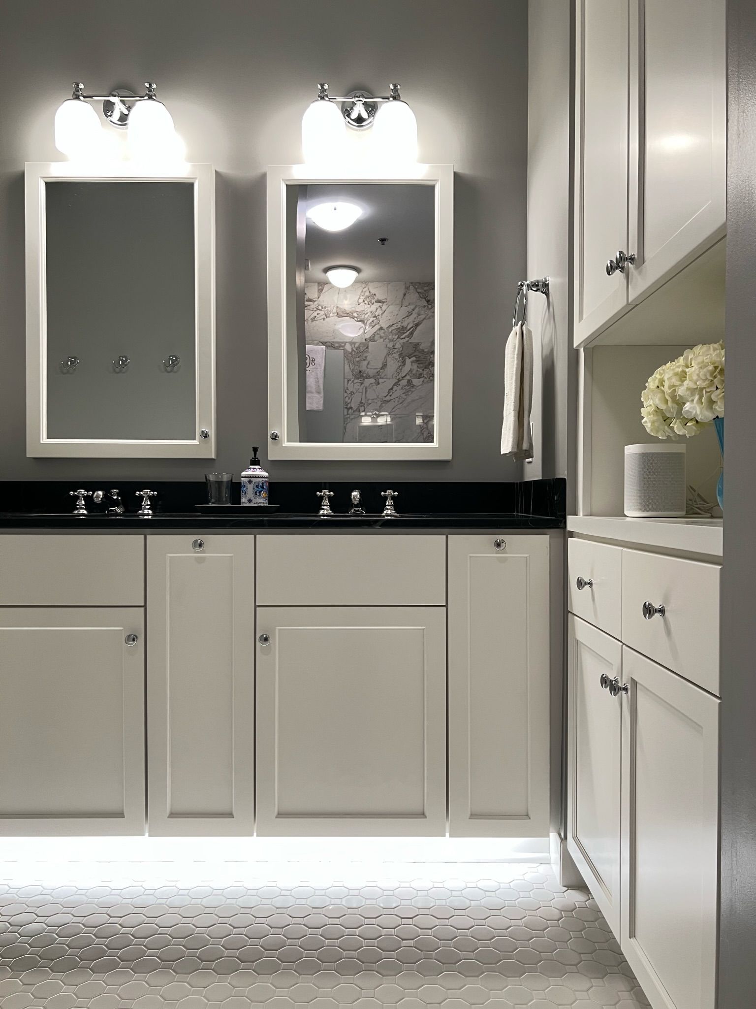 Bathroom with white cabinets, black countertop, two mirrors, and a white hexagonal tile floor.