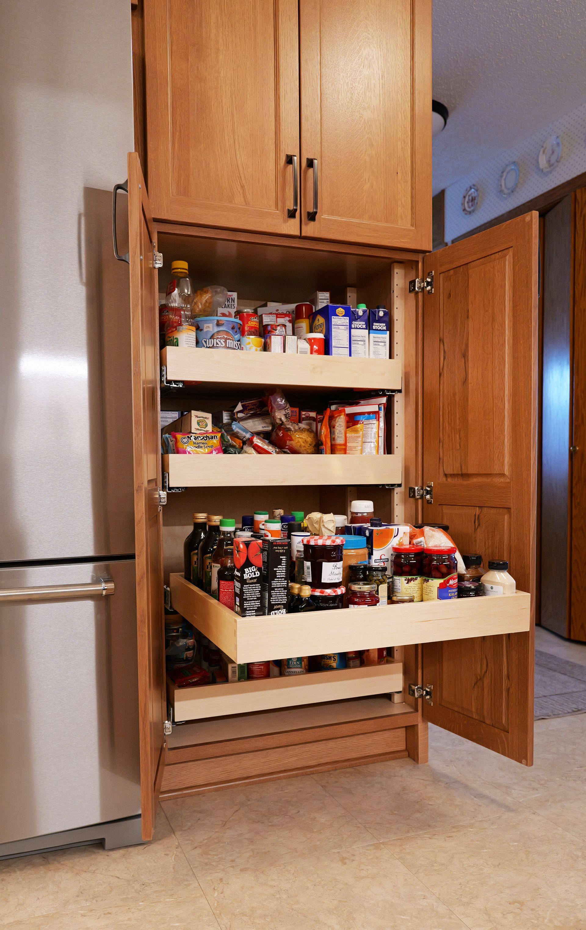 A wooden pantry cabinet with pull-out shelves filled with various food items.  Next to a refrigerator.