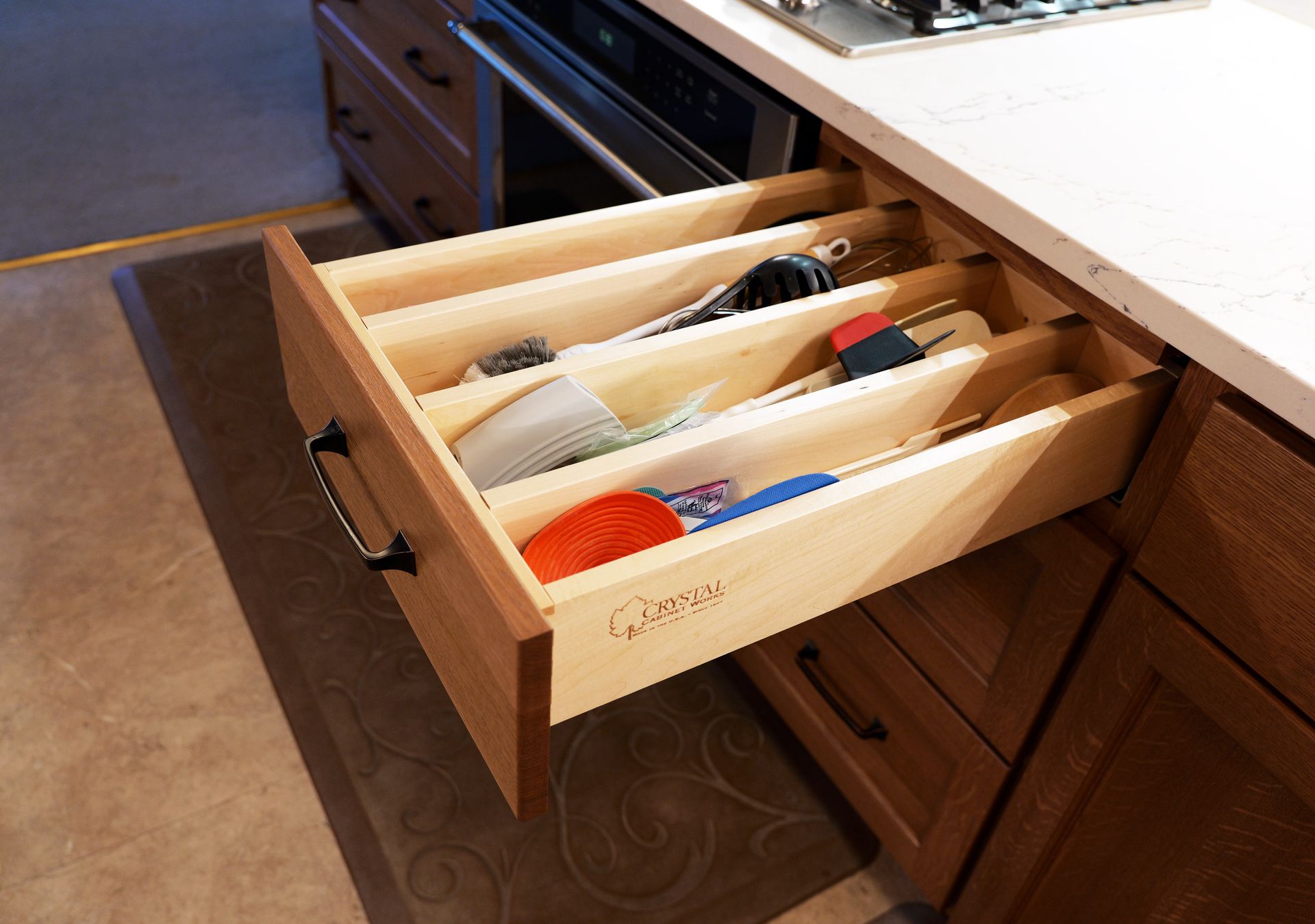 Open kitchen drawer with dividers, holding utensils; wooden cabinets and countertop.