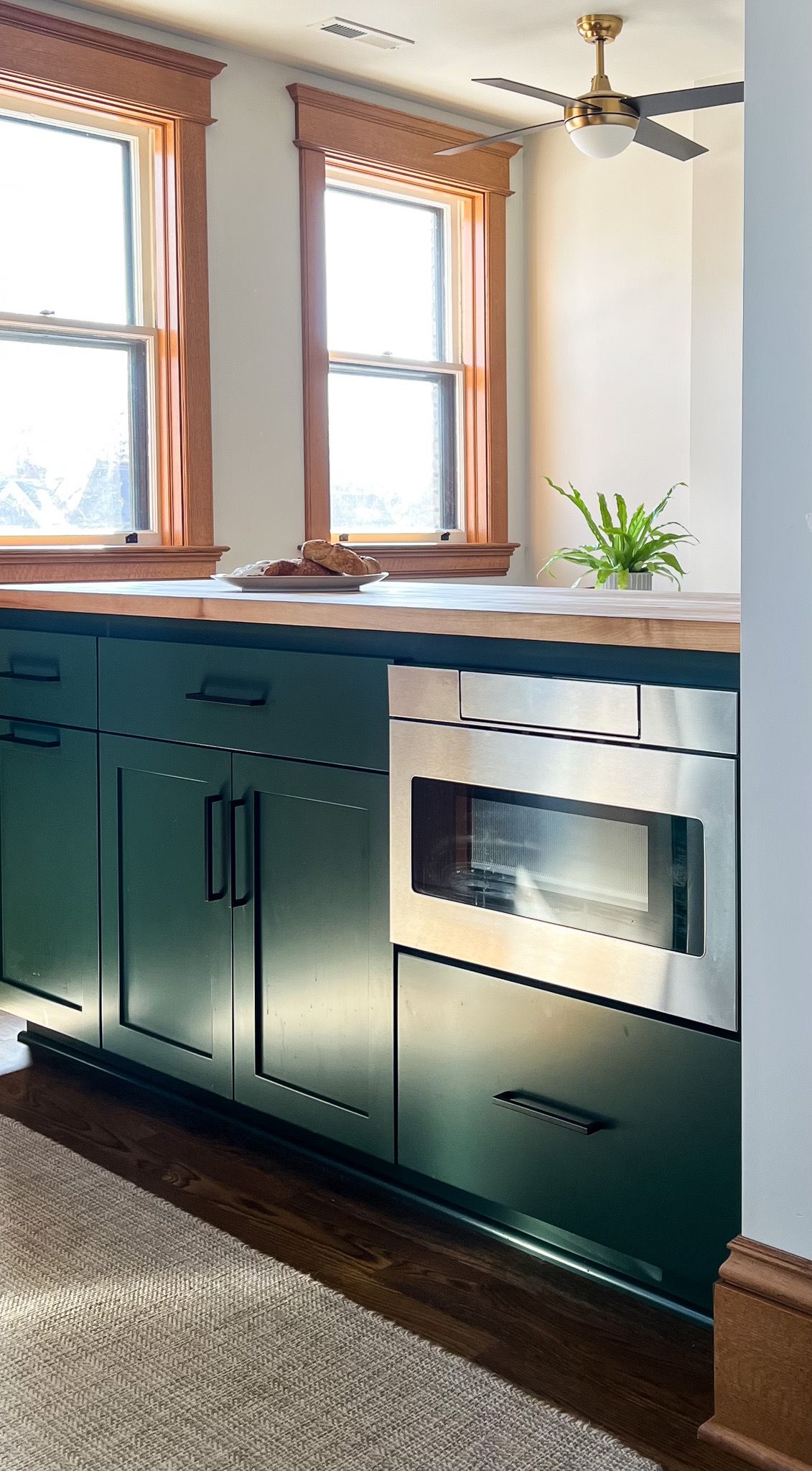 Green kitchen cabinets with stainless steel microwave, wood countertop, and windows with wooden frames.