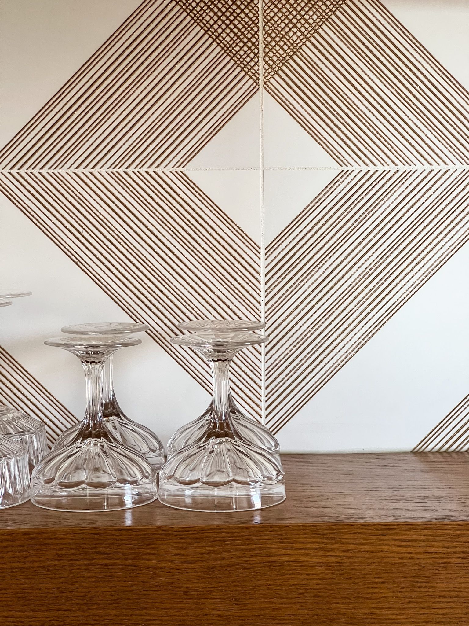 Wine glasses on a wooden shelf, against a geometric diamond-patterned tile wall with brown and white lines.