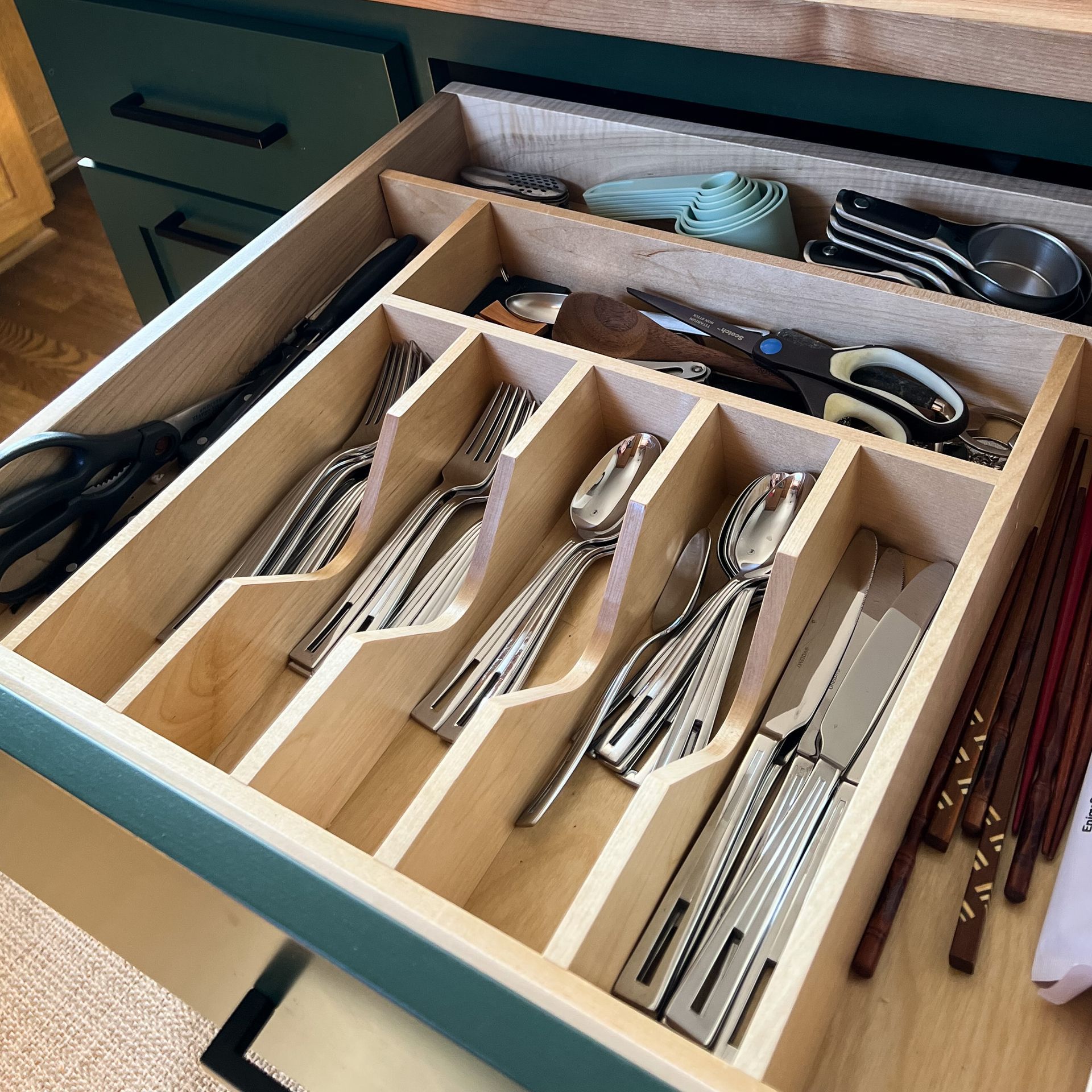 Open drawer with silverware organizer; forks, spoons, knives in compartments. Other kitchen tools visible above.