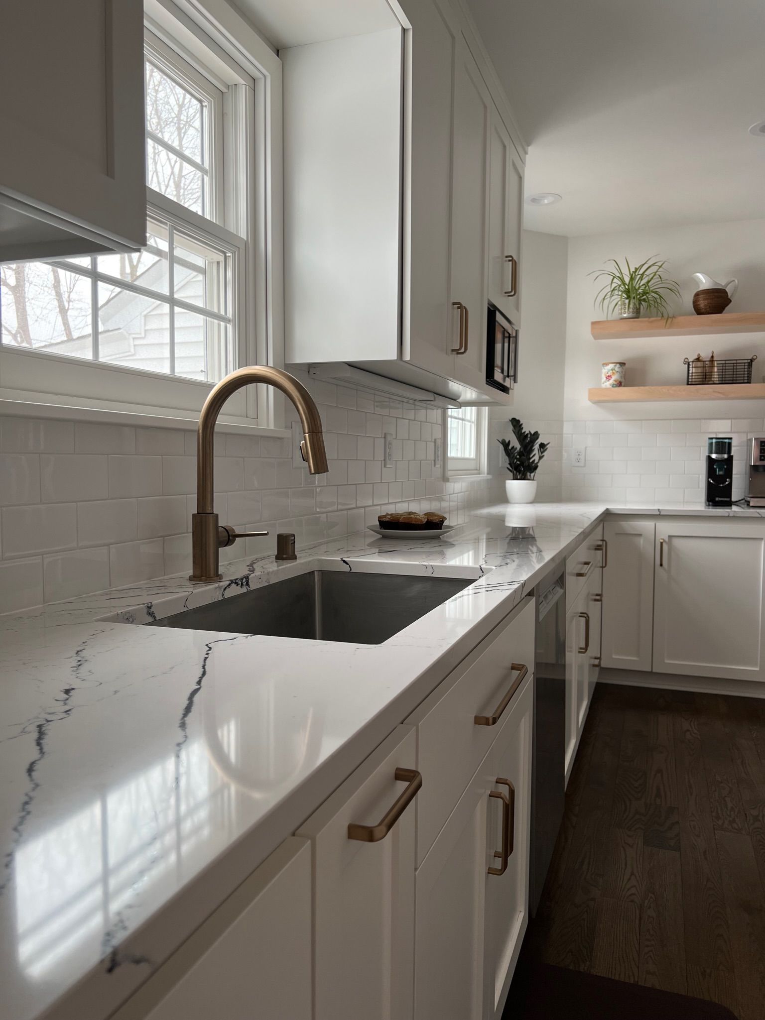 White kitchen with marble countertops, gold faucet, and floating shelves with plants.