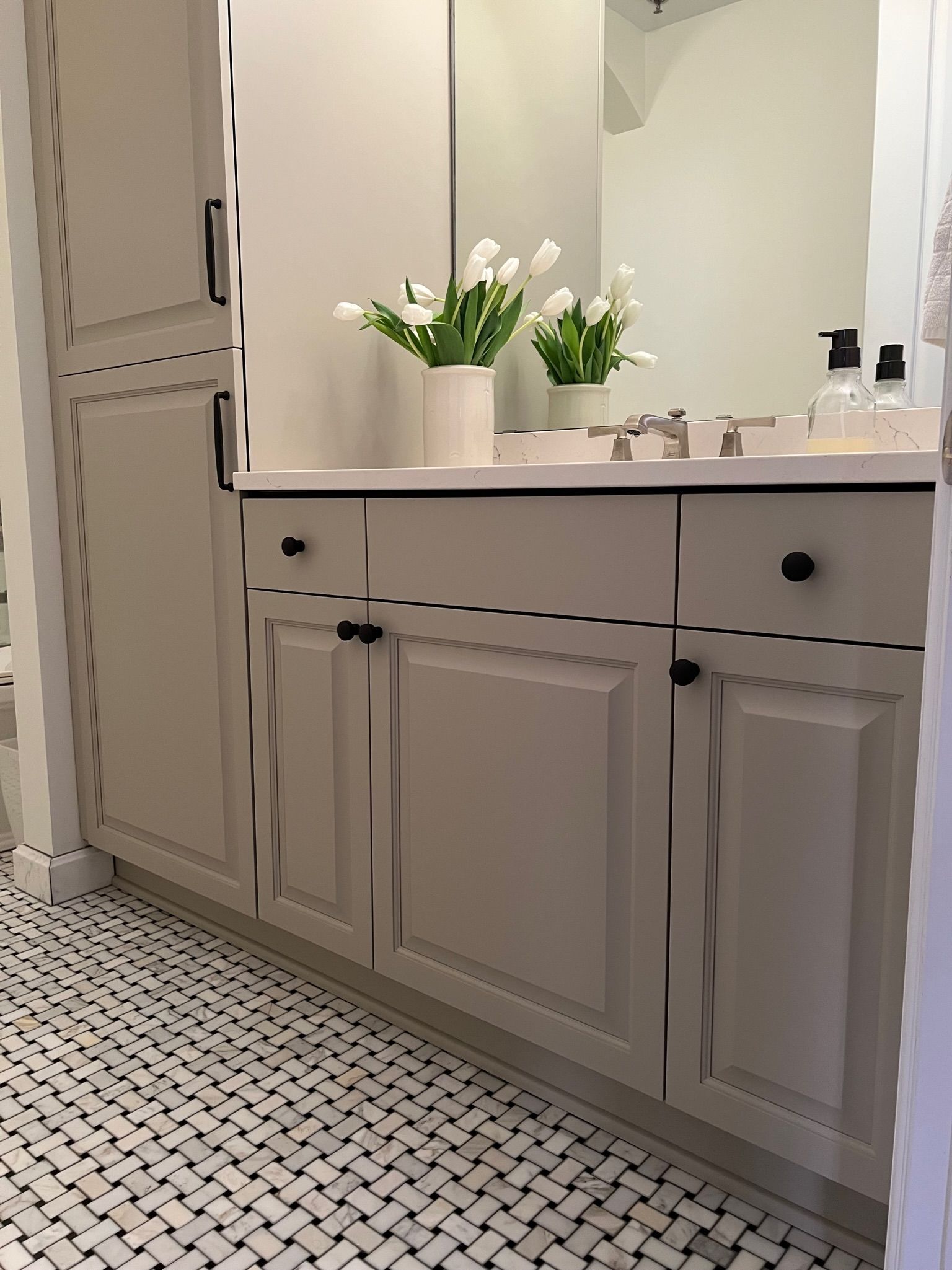 White bathroom vanity with black hardware, next to tall storage cabinet. White tulips in vase. Black and white tiled floor.